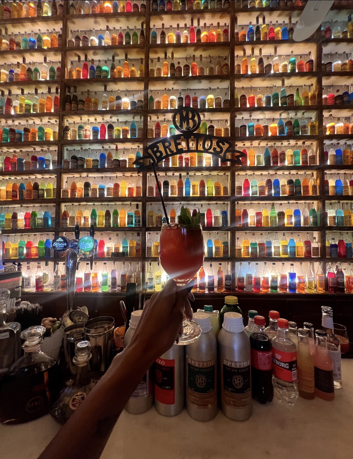 A cocktail held up in front of the colorful bottle wall at Brettos bar in Athens, Greece
