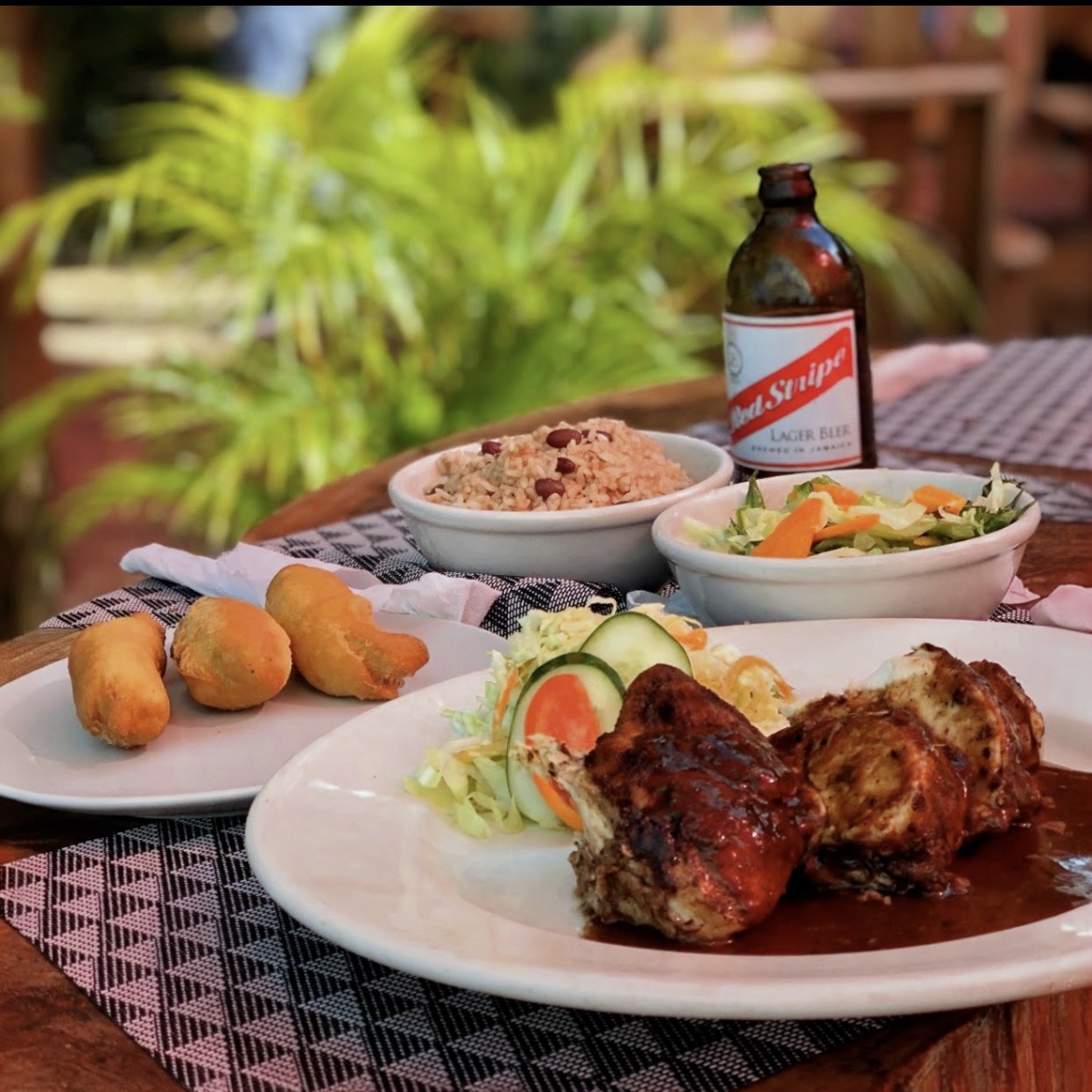 A plate of jerk chicken with rice and peas, coleslaw, fried dumplings, and a Red Stripe beer on a Jamaican restaurant table